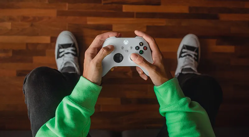 Persoon zit ontspannen op een zwarte bank, spelcontroller in handen. Moderne woonkamer met planten, groot raam en muziekinstrumenten op de achtergrond.