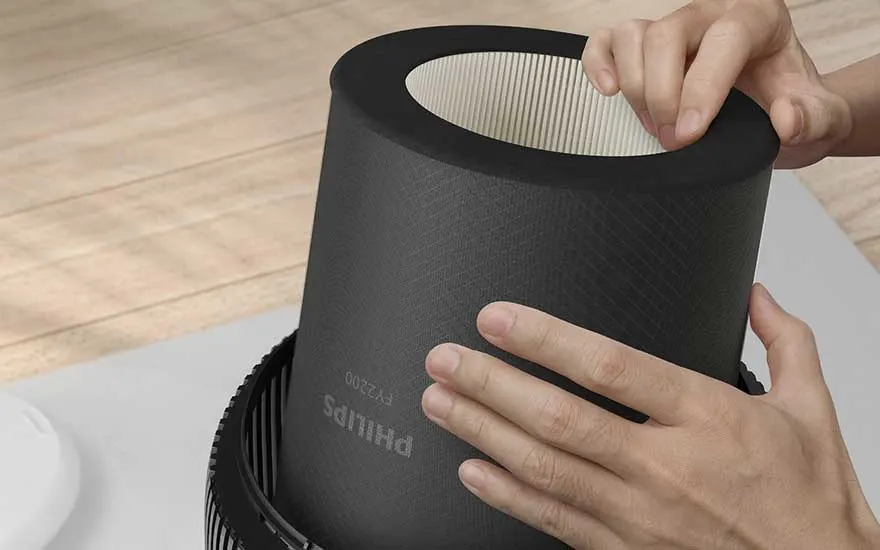 Close-up van handen die een cilindrische Philips-filter in een luchtreiniger plaatsen, boven een houten vloer in een lichte kamer.