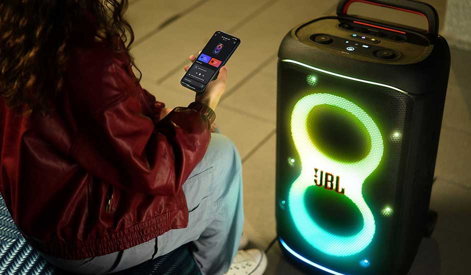 Persoon in rode jas bedient via smartphone een grote JBL-partyspeaker, waarvan de kleurrijke lichtcontouren helder oplichten op de stoep.