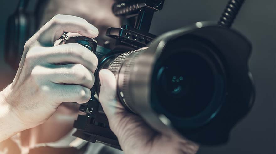 Een persoon bedient een zwarte camera met een grote lens en een microfoon, tegen een vage achtergrond.
