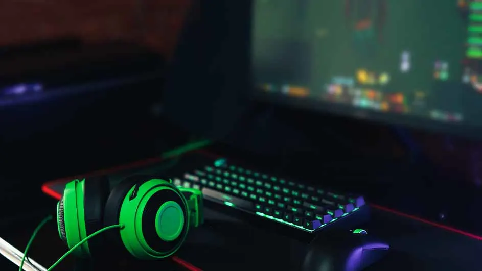 Groene gaming headset naast een toetsenbord met groene verlichting, een muis en een monitor op een donkere tafel.