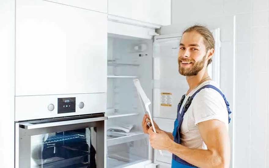 A technician in blue overalls stands beside a modern built-in oven and an open fridge, reading instructions in a bright white kitchen. No brand visible.