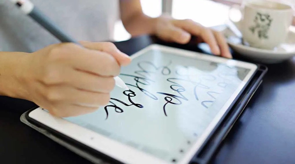 Hands use a stylus on a tablet displaying handwritten words. A mug sits out of focus in the background.