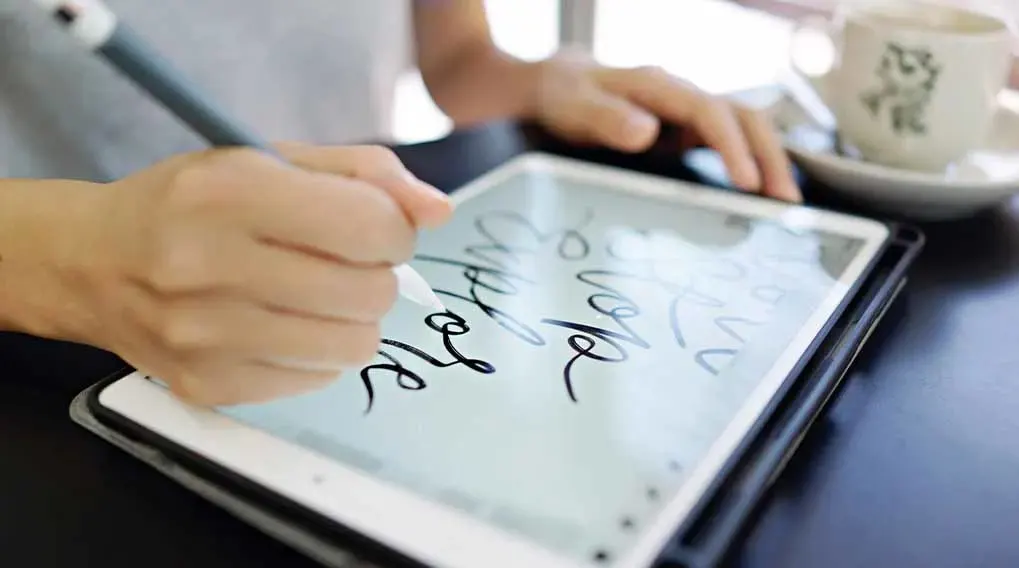 Hand schrijft met een stylus op een tablet; koffiekopje op de achtergrond.
