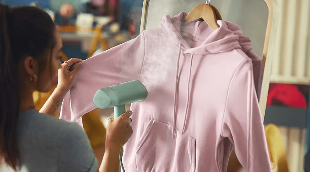 Een vrouw stoomt een roze hoodie op een houten hanger, waarbij stoom uit een groen stoomapparaat komt. Op de achtergrond hangt kleding aan een rek.
