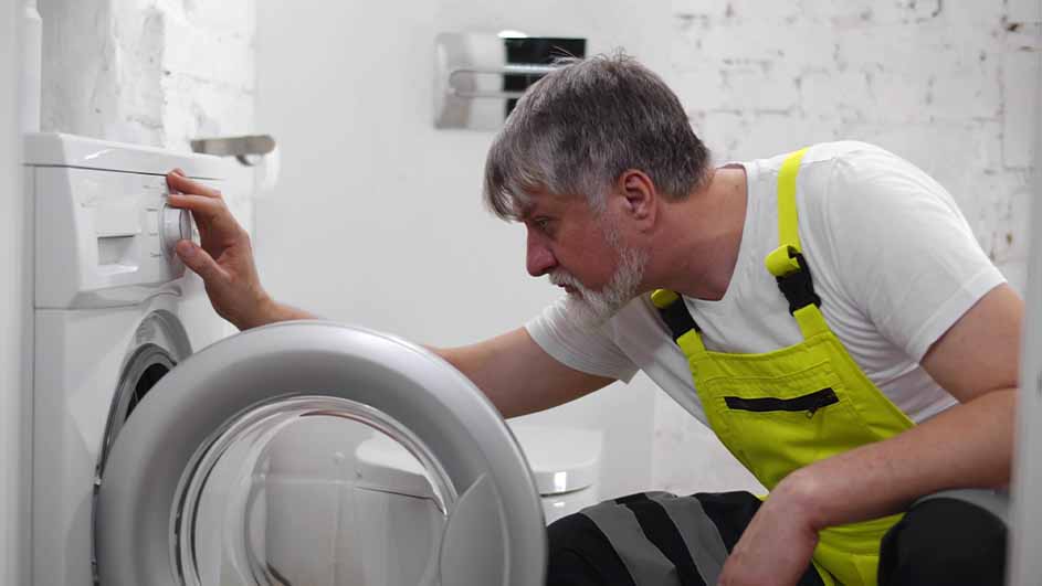 A man in a yellow overall adjusts a white washing machine. The machine has a silver door. He is in a white room with brick details.