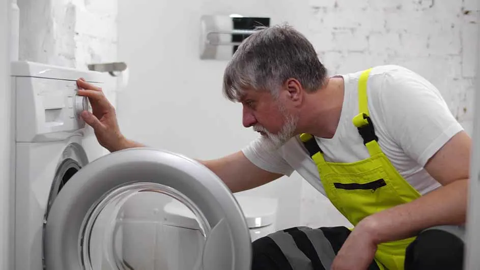 A man in a yellow overall adjusts a white washing machine. The machine has a silver door. He is in a white room with brick details.