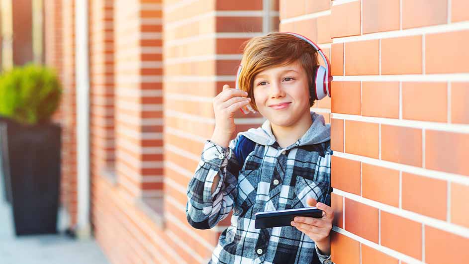 A person with headphones holds a phone, leaning against a brick wall. A green plant is in a black pot in the background.