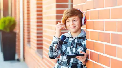 Een jongen met een telefoon en een koptelefoon staat naast een bakstenen muur.