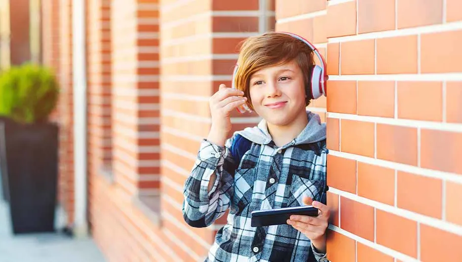 Een jongen met een telefoon en een koptelefoon staat naast een bakstenen muur.