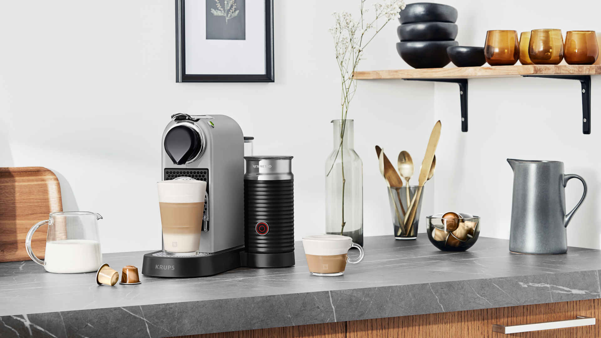 Grijze keuken met Nespresso machine, melkopschuimer en kopjes koffie. Achtergrond toont planken met kommen en glazen.