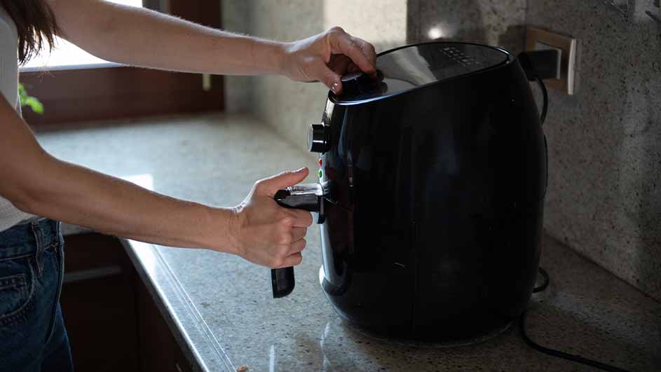 Een persoon bedient een zwarte airfryer op een aanrecht, met een stopcontact zichtbaar op de achtergrond.