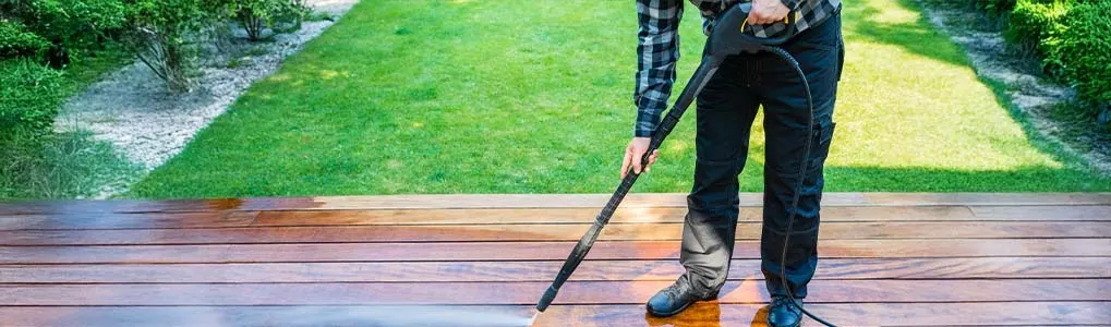 A man is cleaning a wooden terrace with a Kärcher pressure washer, making the surface look bright and fresh in a green garden.