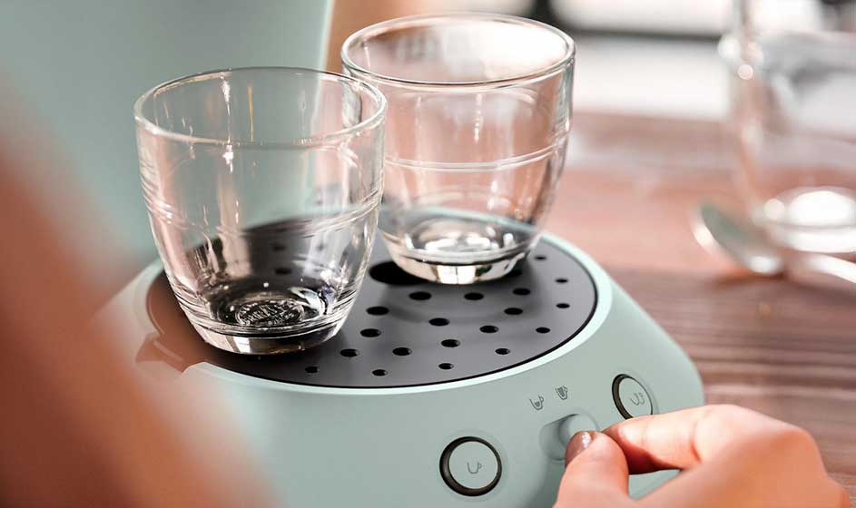 Twee glazen kopjes staan op een lichtgroene koffiemachine, terwijl een hand een knop bedient op een houten tafelblad bij het raam.