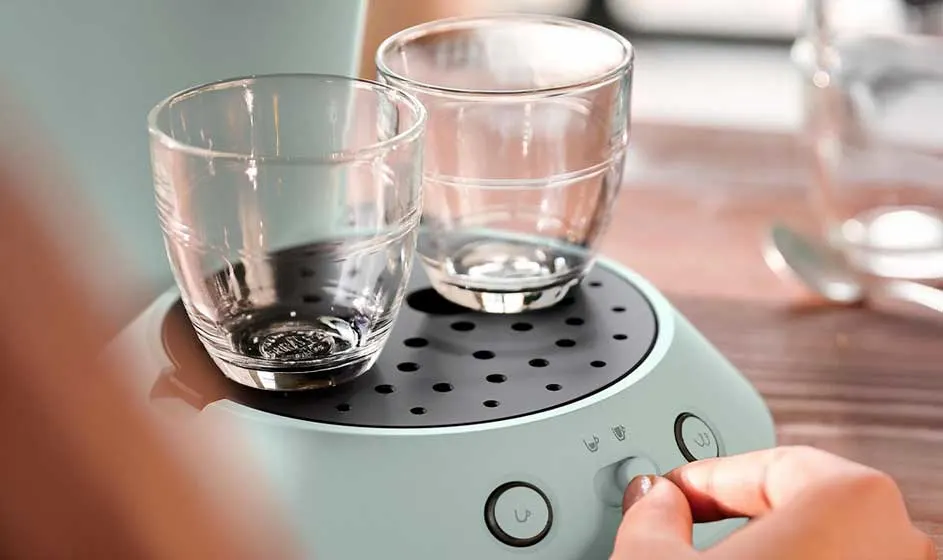Twee glazen kopjes staan op een lichtgroene koffiemachine, terwijl een hand een knop bedient op een houten tafelblad bij het raam.