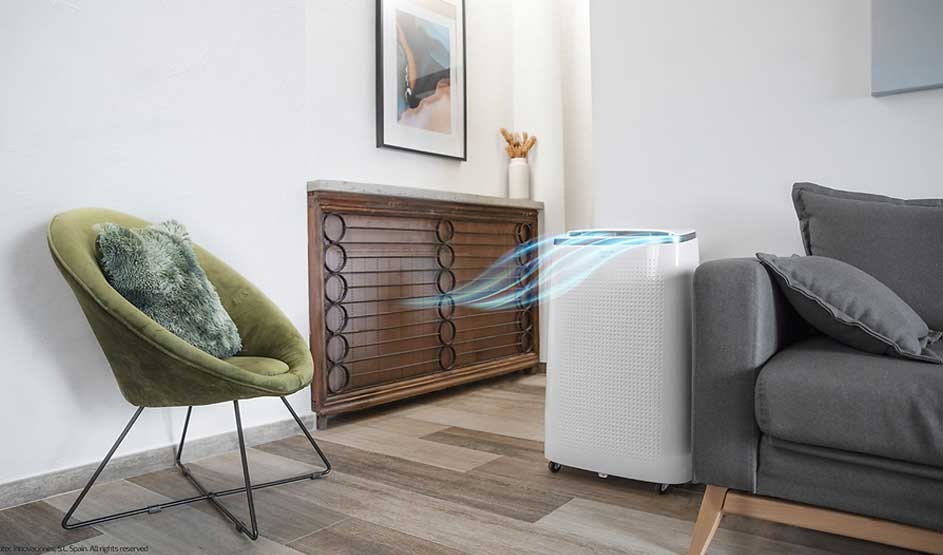 A portable air conditioner from the brand Cecotec is placed beside a grey sofa, gently blowing cool air, in a tidy and cosy living room.