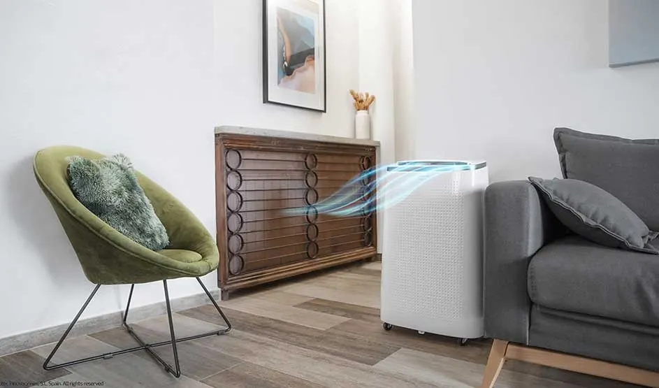 A portable air conditioner from the brand Cecotec is placed beside a grey sofa, gently blowing cool air, in a tidy and cosy living room.