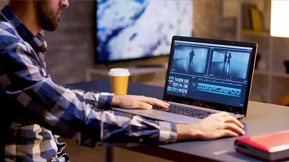 Een man bewerkt video's op een laptop aan een bureau, met een koffiebeker en een rode powerbank ernaast. Op de achtergrond staat een tv.