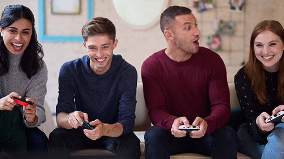 Vier lachende mensen zitten op een bank en spelen videospelletjes met controllers. Er staat een blauw omlijst schilderij aan de muur.