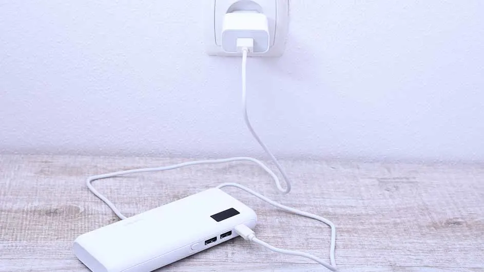 Een witte powerbank ligt op een houten oppervlak en is verbonden met een witte oplader in een stopcontact aan de muur.