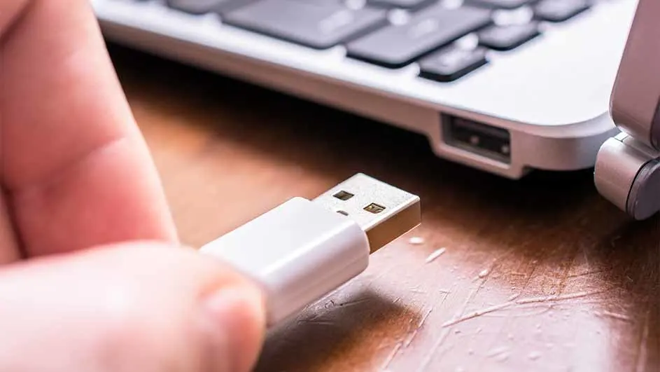 Hand houdt een witte USB-stick vast voor een laptop op een houten bureau.