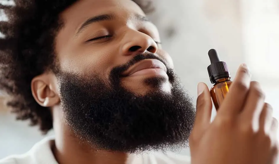 A smiling man with a full beard holds a bottle of beard oil and seems pleased as he enjoys its scent.