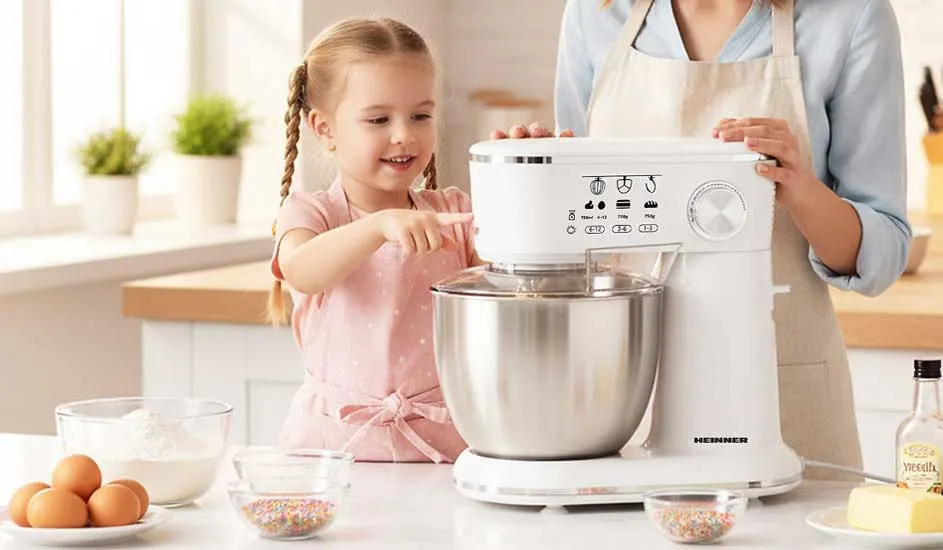 Een meisje in roze schort bakt samen met haar moeder, terwijl een witte keukenmachine beslag klopt tussen eieren, bloem en kleurrijke sprinkles.