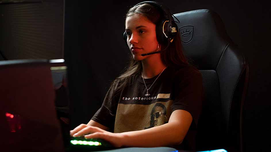 Een vrouw met een headset zit voor een computer, spelend. Ze draagt een zwart T-shirt en zit in een zwarte stoel.
