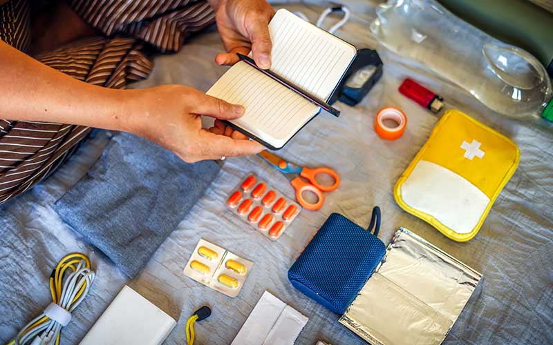 Hands place a small notebook among first-aid items, medications, and emergency supplies on a blanket.