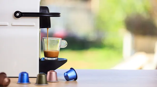 Espresso wordt in een glazen kopje gegoten door een witte machine. Verschillende capsules staan op een tafel voor een groene achtergrond.