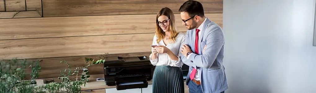 Twee collega’s staan lachend naast een printer in een kantooromgeving, terwijl de vrouw iets op haar smartphone laat zien aan de man in pak.