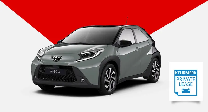 Een grijze Toyota Aygo X staat voor een rode en witte achtergrond, met een blauw keurmerk rechtsonder.