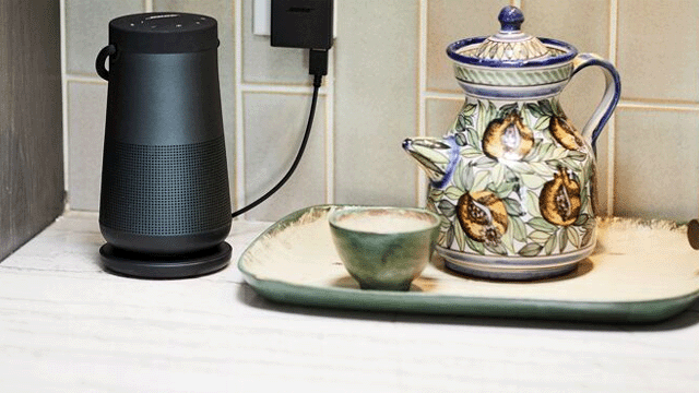 Een zwarte speaker naast een decoratieve theepot en een groene kop op een dienblad. Een oplader zit in het stopcontact.