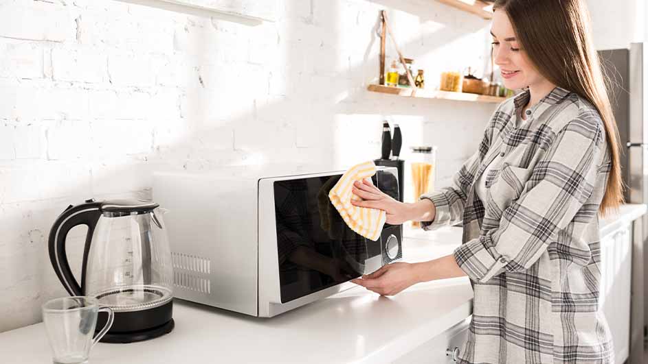 Een vrouw maakt een witte magnetron schoon met een geel-witte doek in een keuken, met een waterkoker en een mok in de buurt.