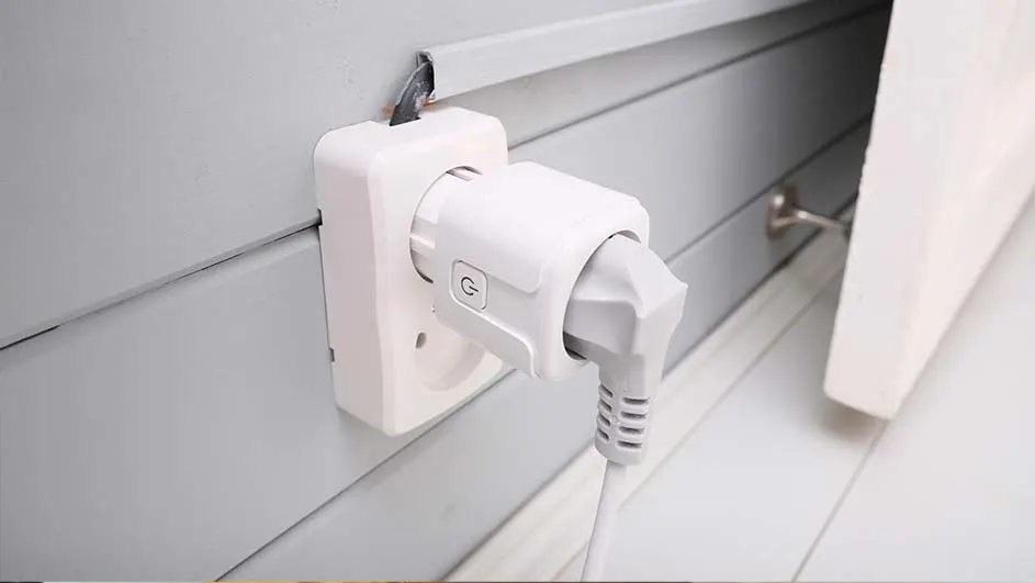 Witte stekker in stopcontact aan grijze wand met plint. Het snoer loopt naar beneden.