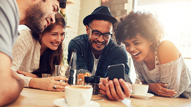 Groep vrienden lacht en kijkt naar een mobiele telefoon aan een tafel in een café met koffiekopjes en een waterfles.