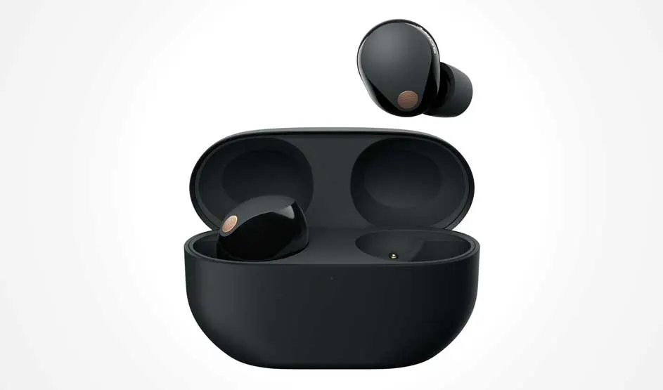 A black charging case sits open; one earbud rests inside and another hovers above, with small copper-toned details on a pale backdrop.