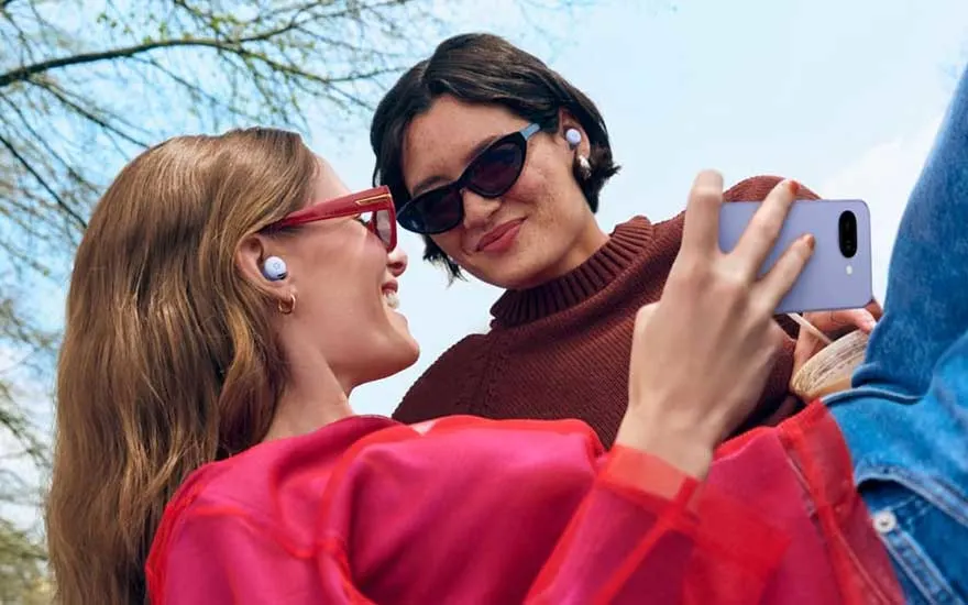 Twee mensen genieten samen buiten, dragen draadloze Pixel Buds oortjes en kijken lachend naar een smartphone in hun hand.