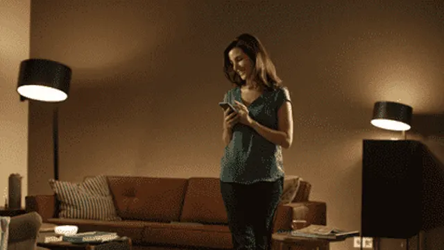 Vrouw staat in een woonkamer en kijkt naar een mobiele telefoon. Er staan lampen en een bruine bank in de kamer.