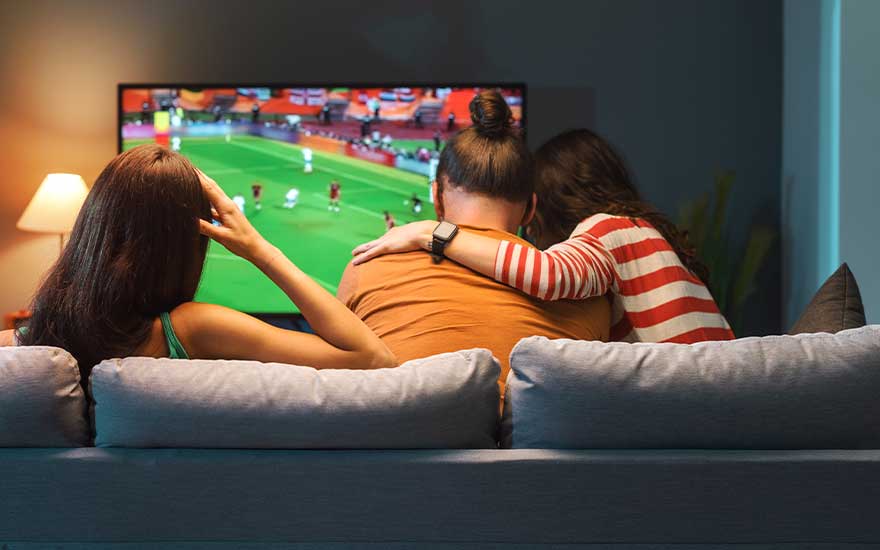 Drie mensen zitten samen op de bank terwijl ze naar een voetbalwedstrijd op tv kijken, genietend van de sfeer thuis.