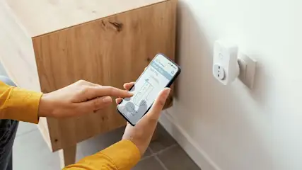 Een persoon bedient een smartphone met een app voor een slimme stekker, die in een stopcontact aan een witte muur zit.