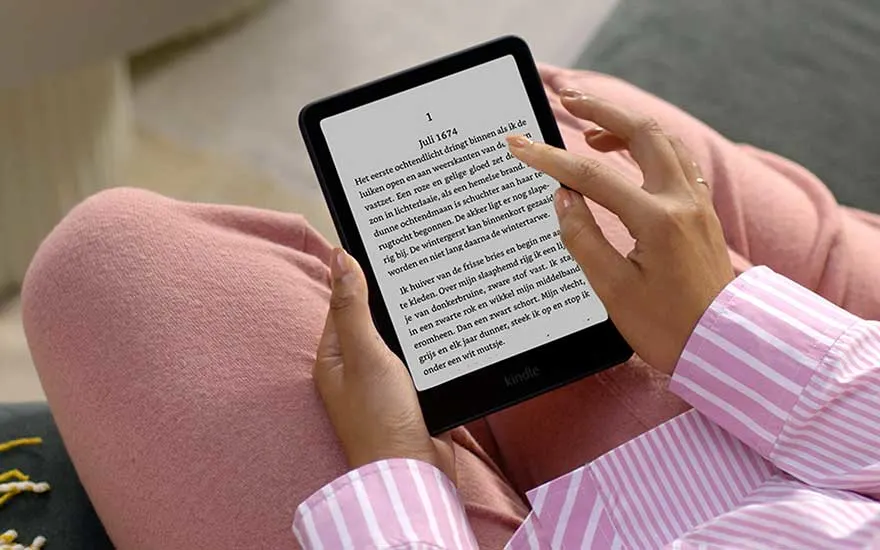 Someone in pink clothing holds a Kindle and calmly reads a Dutch text while sitting in a relaxed, comfortable position.