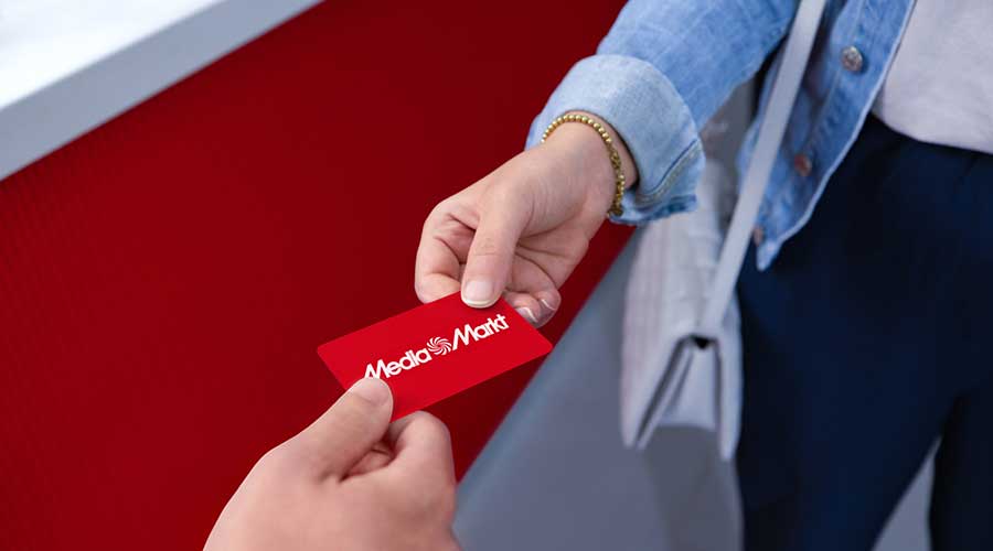Hand in denim blouse receives red MediaMarkt gift card against a red background.