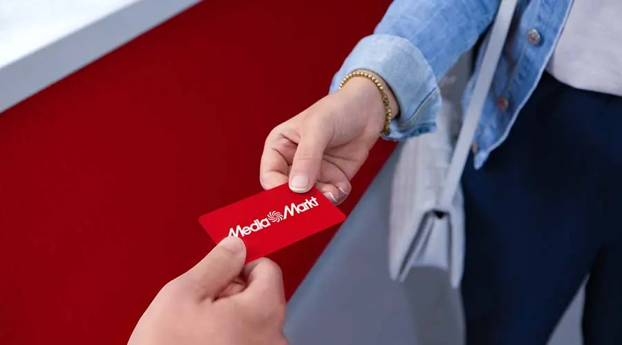 Hand in spijkerblouse ontvangt rode MediaMarkt cadeaukaart tegen rode achtergrond.