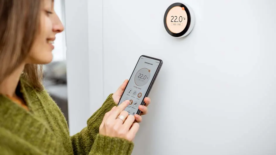Een vrouw in een groene trui bedient een witte thermostaat met een telefoon. De temperatuur is 22 graden.