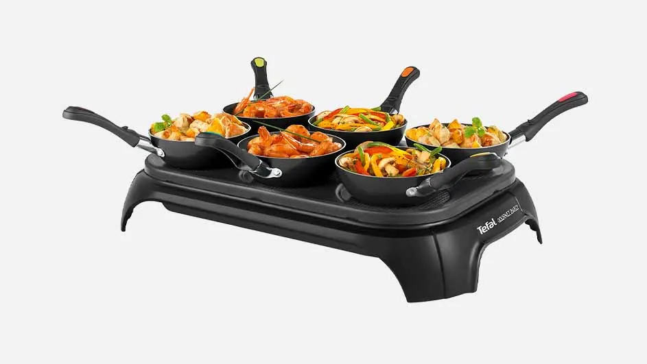 Zes zwarte pannetjes met diverse gerechten op een zwarte elektrische bakplaat tegen een witte achtergrond.