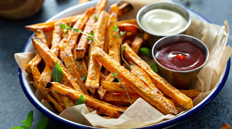Een blauwe schaal gevuld met zoete aardappelfriet, twee dipsausjes en bakpapier op een donkere ondergrond.