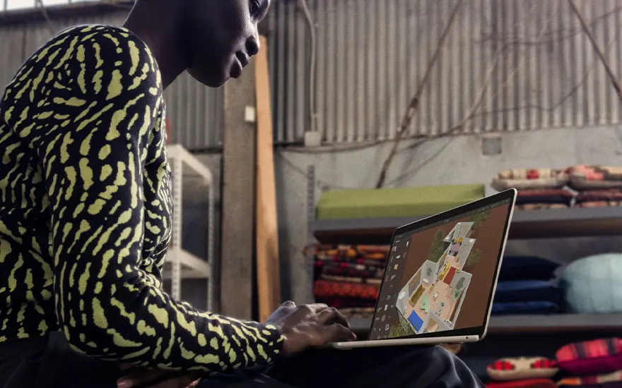 Man in geel-zwart gestreept shirt werkt aan laptop in industriële ruimte, scherm toont plattegrond, magazijn met dozen op achtergrond.