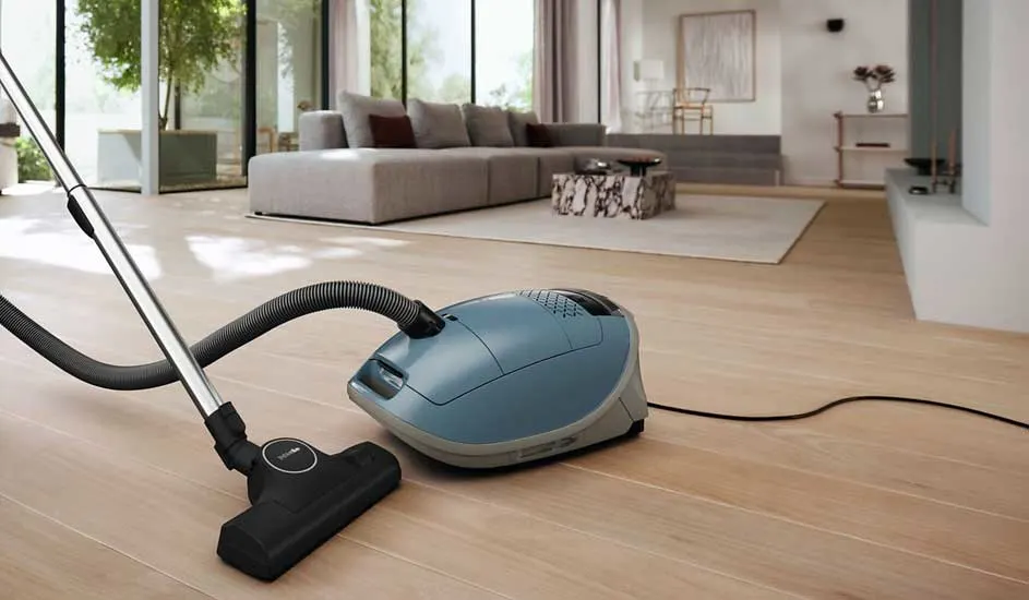 A blue cylinder vacuum rests on pale wooden flooring in a stylish living room with wide windows and a large grey corner sofa.