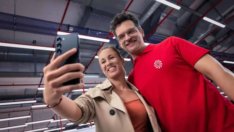 Vrouw en man maken een selfie met een zwarte smartphone in een industrieel ogende ruimte met buizen en lampen aan het plafond.